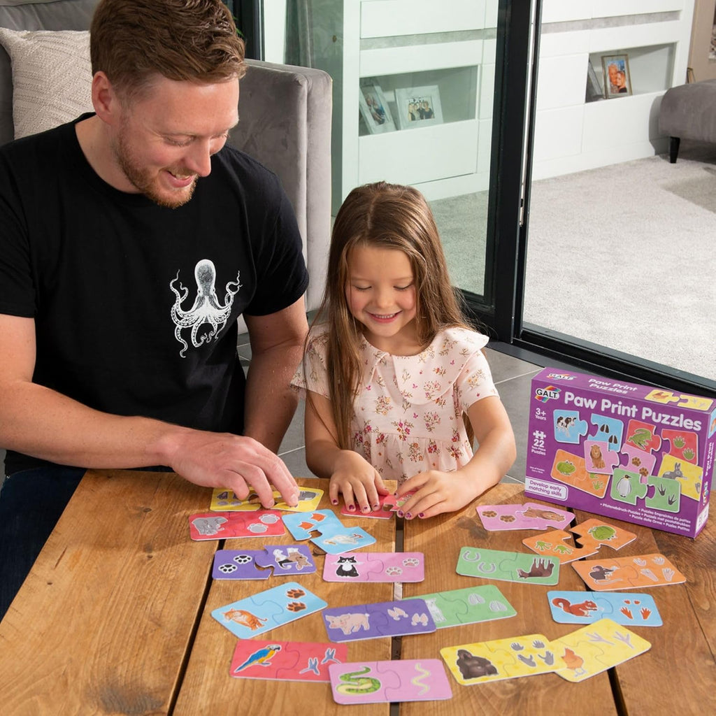 Galt Puzzle - Paw Prints. Man and young girl playing with puzzle cards on a wooden table, with a 'Paw Print Puzzles' box in the background.