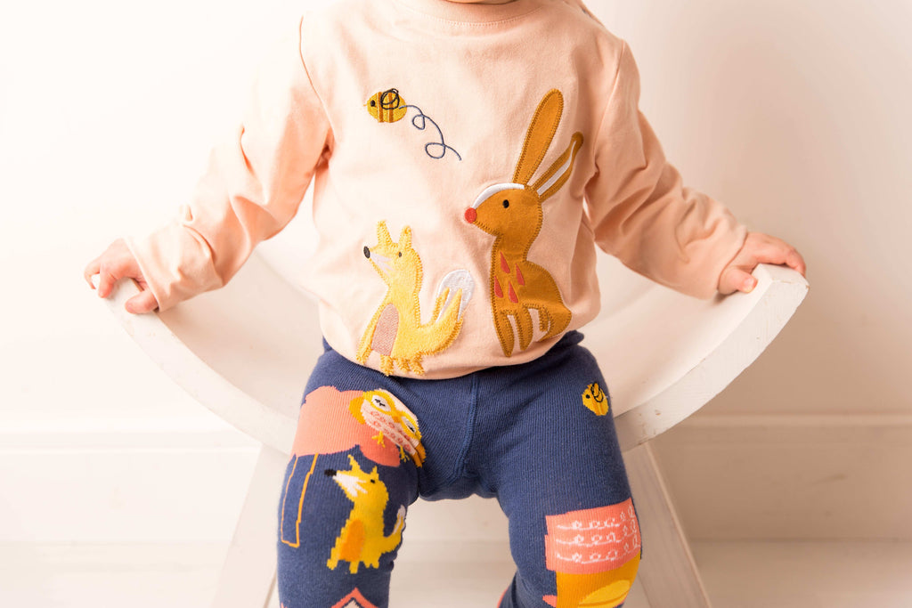 Child wearing a light pink shirt with animal prints and dark blue leggings with colorful designs on a white background. Blade & Rose Meadow Farmyard Top