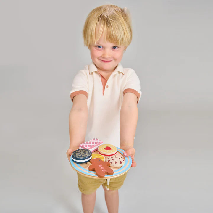Mentari Tea Time Biscuit Plate. Child holding a wooden toy with biscuit shapes against a gray background. Child holding a toy plate with various food items on a plain background
