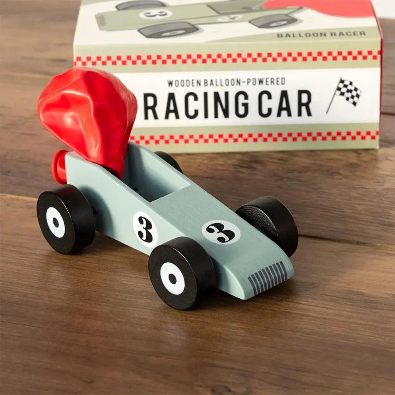 Rex London Wooden Balloon-Powered Racing Car. Wooden balloon-powered racing car with red nose and black wheels on a wooden surface, in front of its packaging.