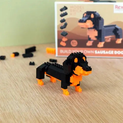 Build Your Own Mini Sausage Dog Brick Kit.T oy sausage dog made of interlocking bricks on a wooden surface with a box in the background.