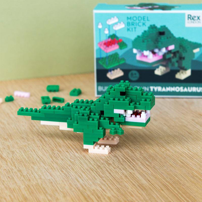 Build Your Own Mini T. Rex Dinosaur Brick Kit. Green dinosaur model brick kit on a wooden surface with packaging in the background.