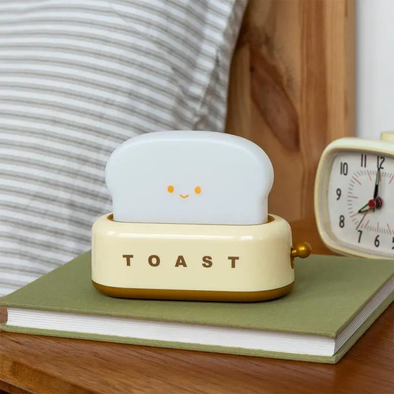 Rex London USB Rechargeable Dimmable Night Light - Kawaii Toaster. Small toaster-shaped night light with a smiley face on top, sitting on a book next to an alarm clock.
