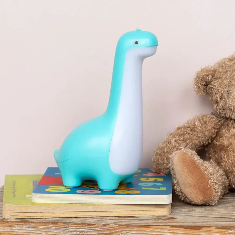 Rex London USB Rechargeable Dimmable Night Light - Dinosaur. Turquoise dinosaur-shaped night light on a stack of books with a teddy bear in the background.