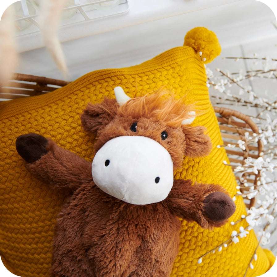 Warmies Highland Cow - Plush cow toy on a yellow knitted pillow with a blurred background