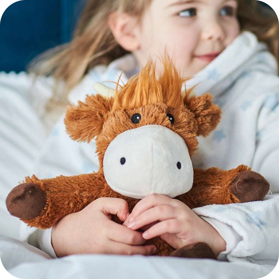 Warmies Highland Cow - Child holding a brown plush highland cow toy with a soft white blanket in the background 