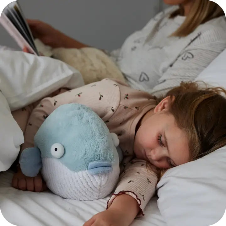 Warmies Pufferfish - Child lying in bed with a plush toy and reading a book, surrounded by soft bedding.