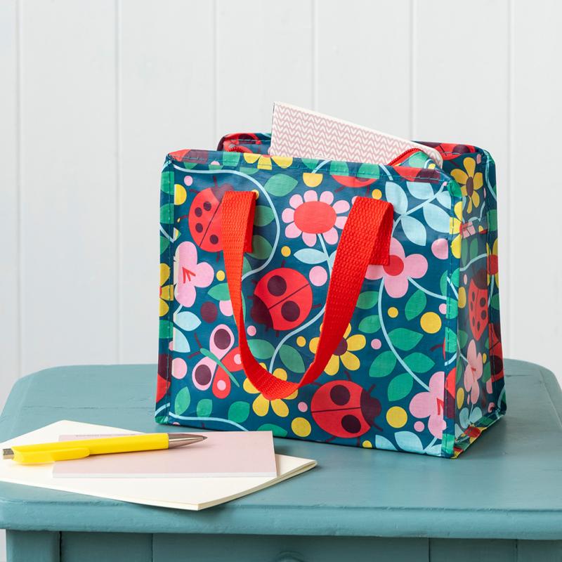 This fabulous Ladybird design Charlotte Bag features a colourful ladybird and flower design on a dark blue background with bright red handles. Say It Baby Gifts